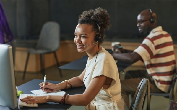 Mulher sentada à mesa de trabalho, com computador e caderno de anotações. Ela usa um fone de ouvido e parece trabalhar com customer service