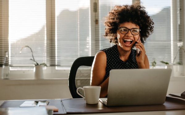 Mulher sorrindo em frente a um notebook enquanto fala ao celular. Transmitindo confiança para um bom relacionamneto com o cliente