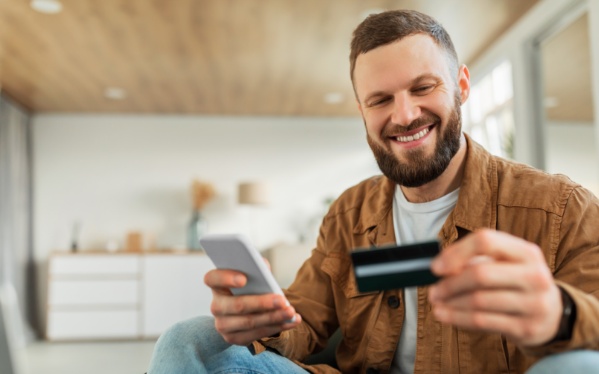 Homem branco com barba sorri ao olhar um cartão de crédito segurado por uma de suas mãos. Na outra está um celular.